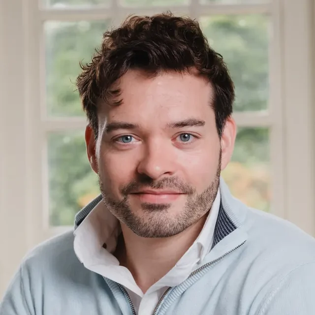 John King, smiling, in a pale blue jumper, sitting by a window