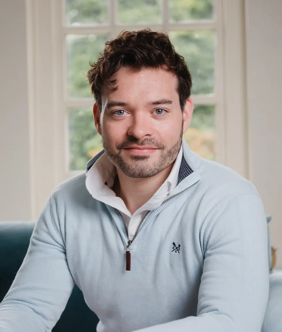 John King, smiling, in a pale blue jumper, sitting by a window